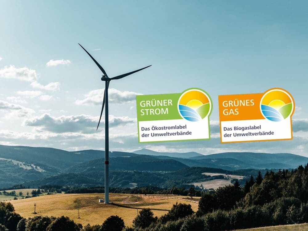 A photo of a wind turbine in front of a sunny, mountainous landscape with forest and fields, with the Grüner Strom Label e.V. next to it.